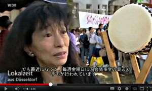 Im Hintergrund Demonstranten und Transparent: »Sayonara…«.